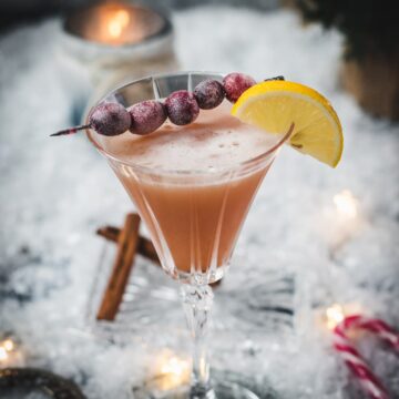 Festive cranberry cinnamon whiskey sour in gorgeous glass, placed on ice ground next to cinnamon sticks and candy canes.