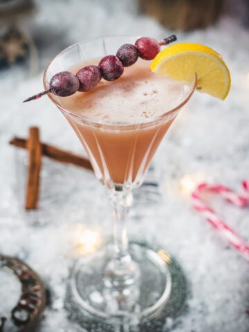 Festive cranberry cinnamon whiskey sour in gorgeous glass, placed on ice ground next to cinnamon sticks and candy canes.