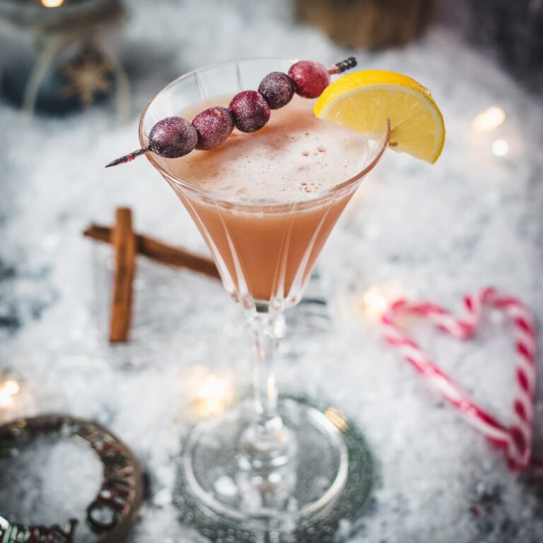 Festive cranberry cinnamon whiskey sour in gorgeous glass, placed on ice ground next to cinnamon sticks and candy canes.