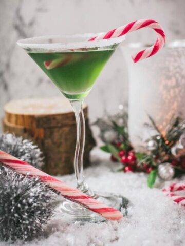 Festive cranberry peppermint martini in sugar rimmed martini glass. Garnished with a candy cane, placed on snowy ground.