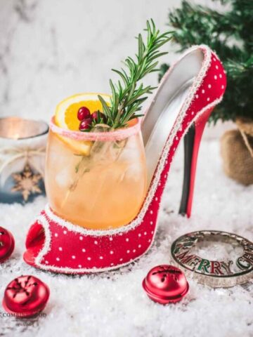 Festive vodka cranberry spritzer, garnished with rosemary twig, orange slice, and cranberries, served in a glass that is placed in a christmassy red high heel.