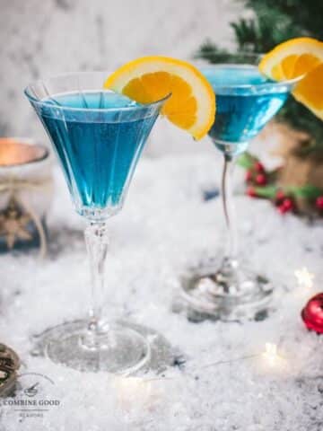 Gorgeous cocktail glasses filled with a stunning blue Christmas cocktail, garnished with an orange slice. Placed on mirrored coasters on snowy ground.