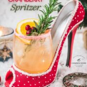 Festive vodka cranberry spritzer, garnished with rosemary twig, orange slice, and cranberries, served in a glass that is placed in a christmassy red high heel.