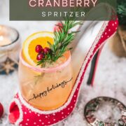 Festive vodka cranberry spritzer, garnished with rosemary twig, orange slice, and cranberries, served in a glass that is placed in a christmassy red high heel.