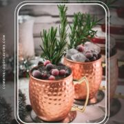 Two copper mugs filled with a delicious cranberry Moscow mule, placed on mirrored coaster and garnished with rosemary twigs and frozen cranberries.