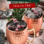 Two copper mugs filled with a delicious cranberry Moscow mule, placed on mirrored coaster and garnished with rosemary twigs and frozen cranberries.