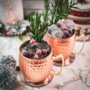 Two copper mugs filled with a delicious cranberry Moscow mule, placed on mirrored coaster and garnished with rosemary twigs and frozen cranberries.