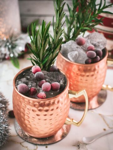 Two copper mugs filled with a delicious cranberry Moscow mule, placed on mirrored coaster and garnished with rosemary twigs and frozen cranberries.