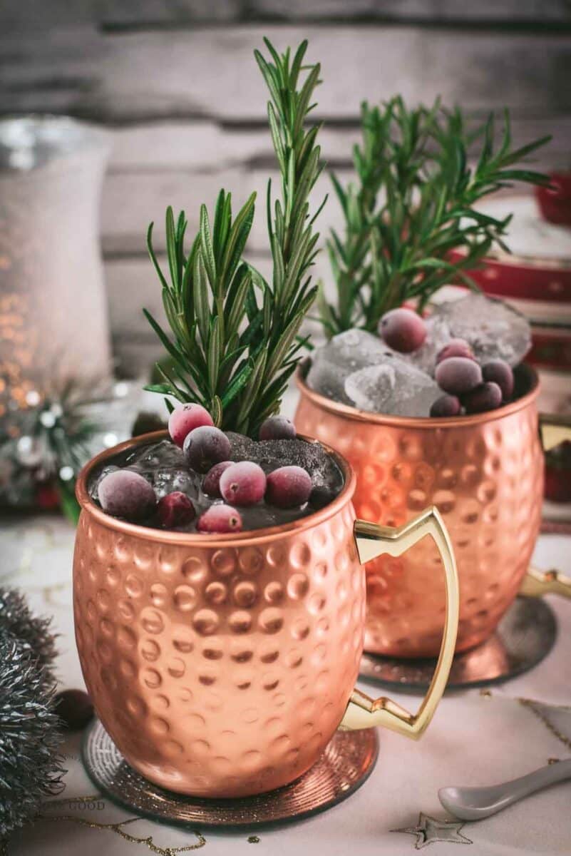 Two copper mugs filled with a delicious cranberry Moscow mule, placed on mirrored coaster and garnished with rosemary twigs and frozen cranberries.