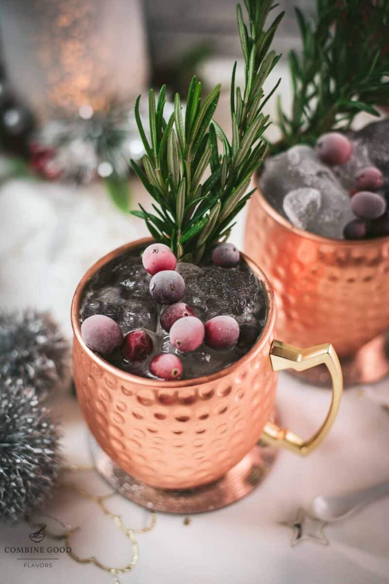 Two copper mugs filled with a delicious cranberry Moscow mule, placed on mirrored coaster and garnished with rosemary twigs and frozen cranberries.