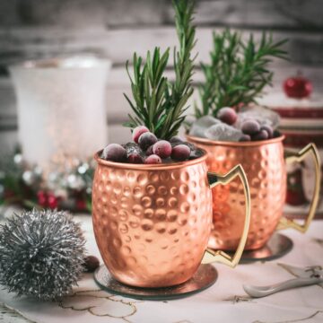 Two copper mugs filled with a delicious cranberry Moscow mule, placed on mirrored coaster and garnished with rosemary twigs and frozen cranberries.