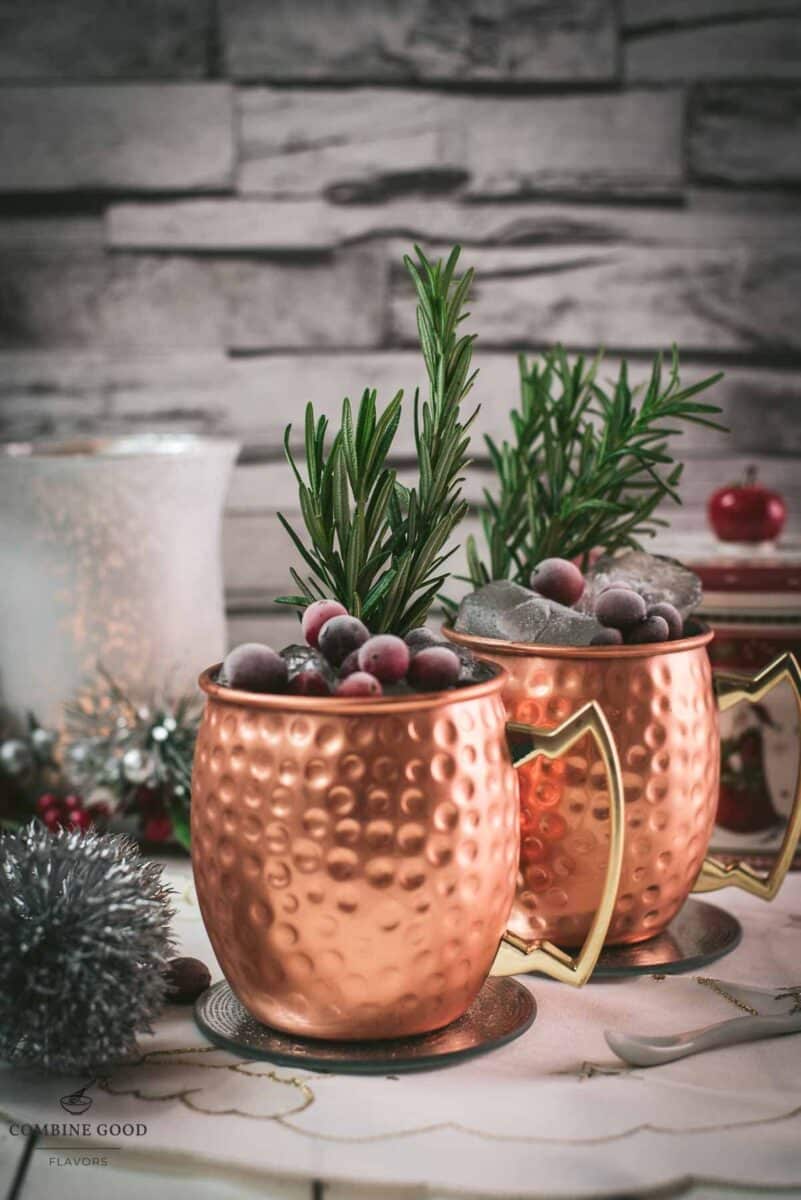 Two copper mugs filled with a delicious cranberry Moscow mule, placed on mirrored coaster and garnished with rosemary twigs and frozen cranberries.