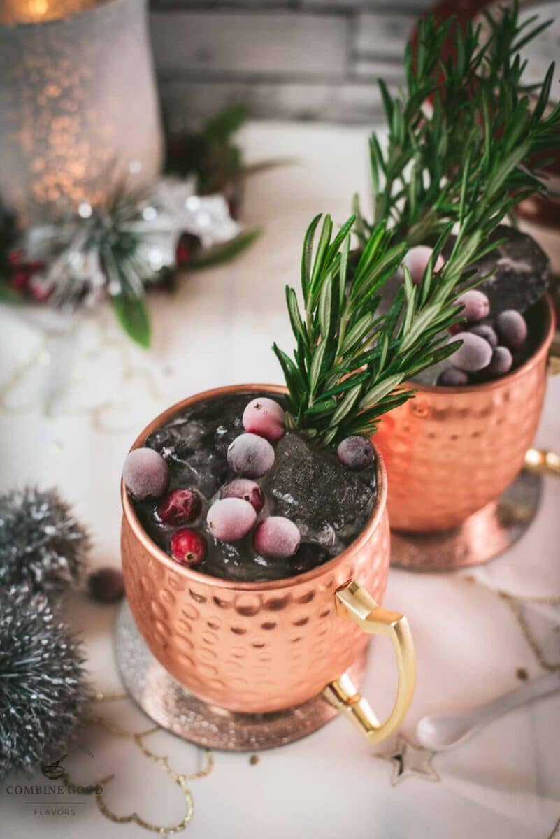 Two copper mugs filled with a delicious cranberry Moscow mule, placed on mirrored coaster and garnished with rosemary twigs and frozen cranberries.