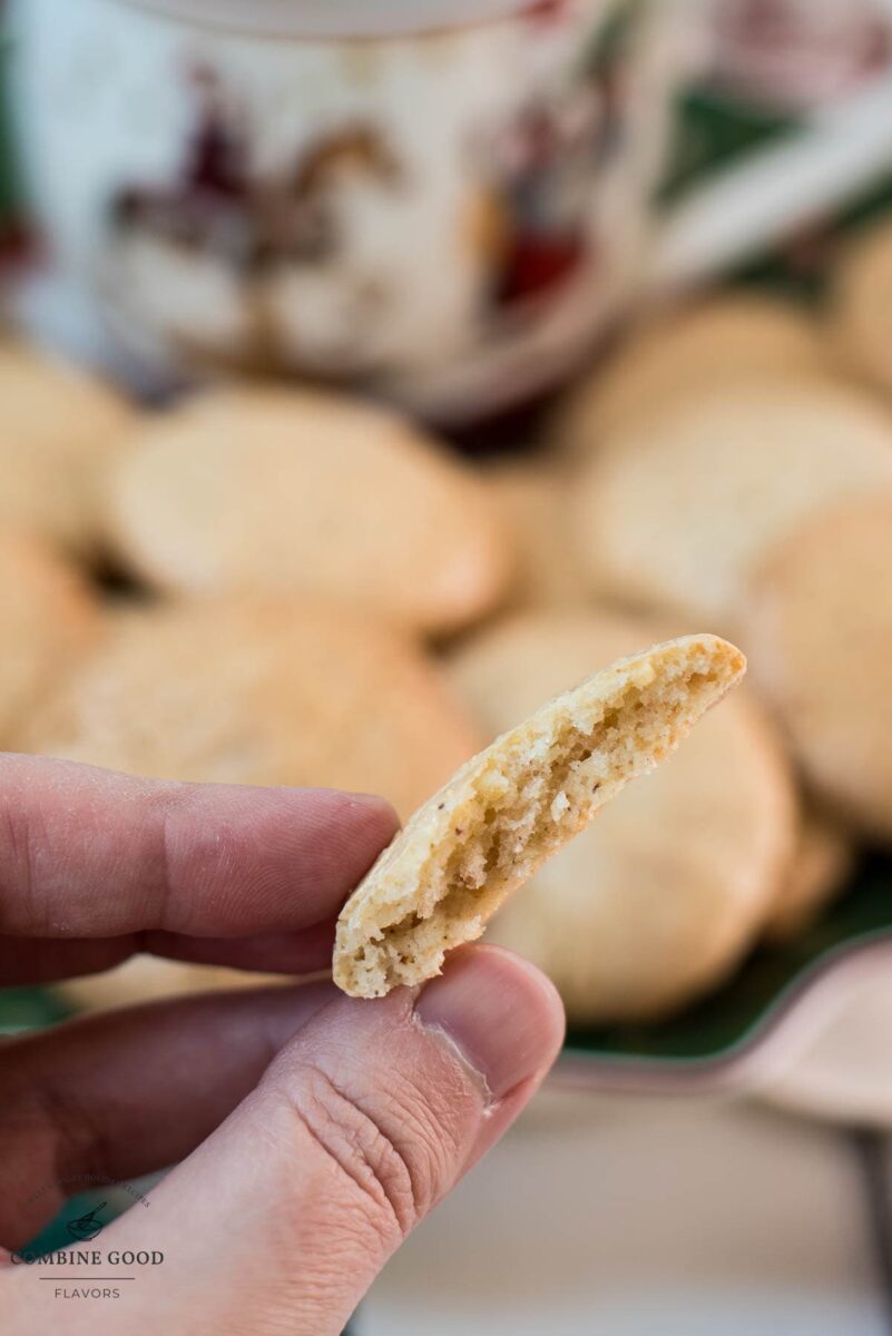 Two fingers holding half a hazelnut meringue cookie in front of a filled cookie plate.