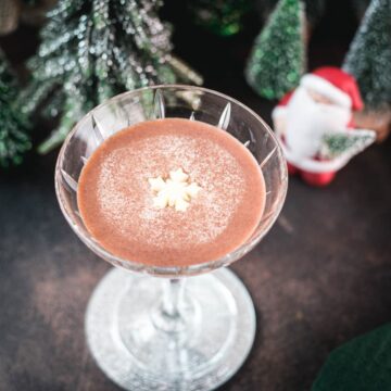 Festive chocolate martini with a white chocolate snowflake garnish, placed on mirrored coaster next to a Santa clause figurine.