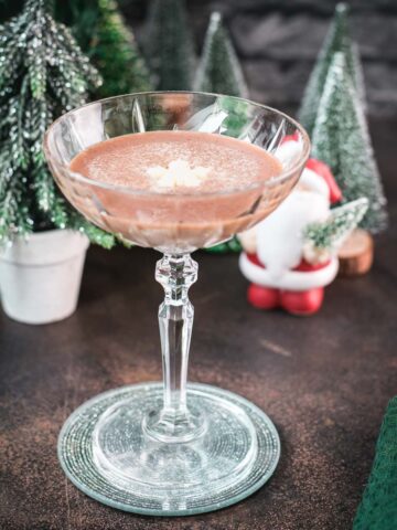 Festive chocolate martini with a white chocolate snowflake garnish, placed on mirrored coaster next to a Santa clause figurine.