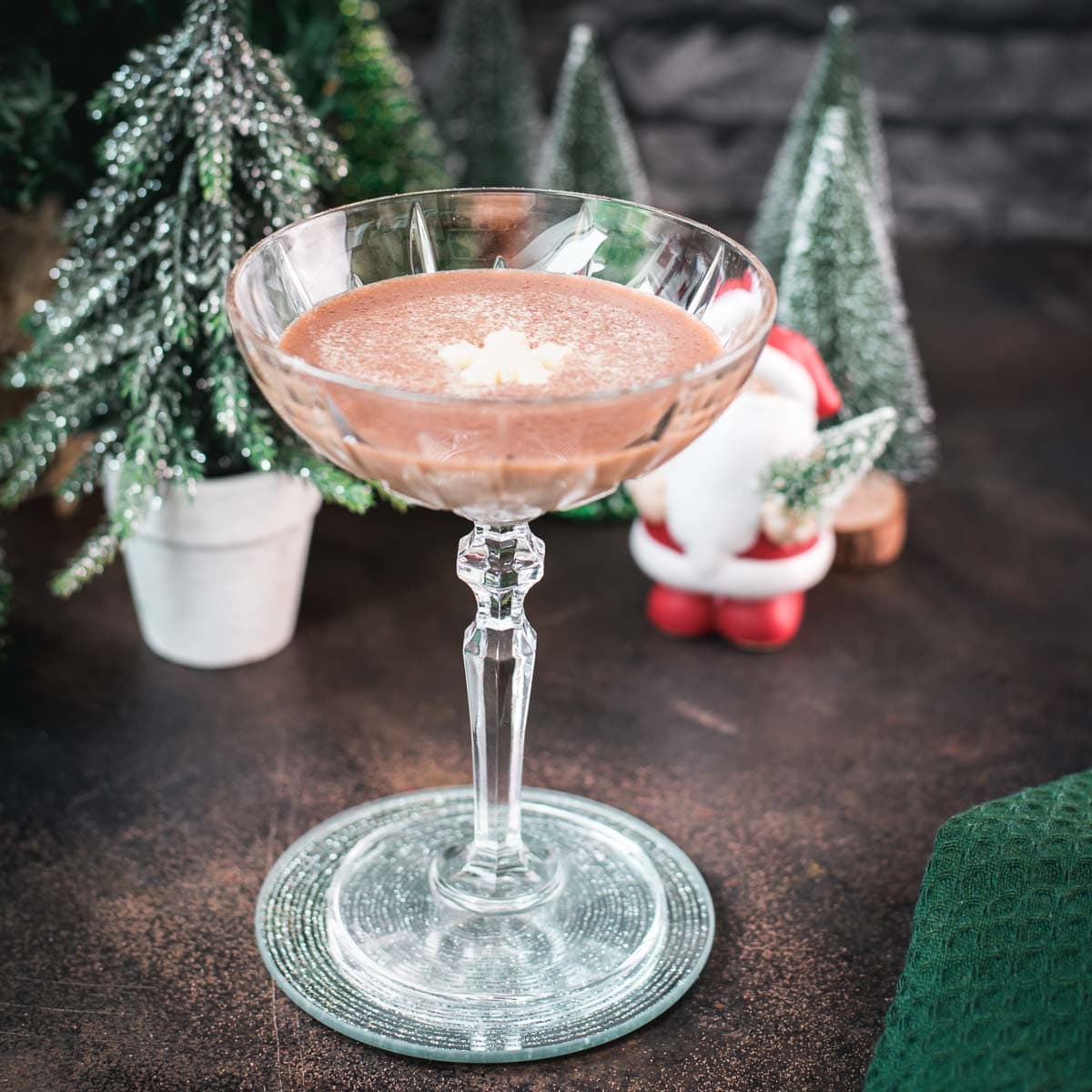 Festive chocolate martini with a white chocolate snowflake garnish, placed on mirrored coaster next to a Santa clause figurine.