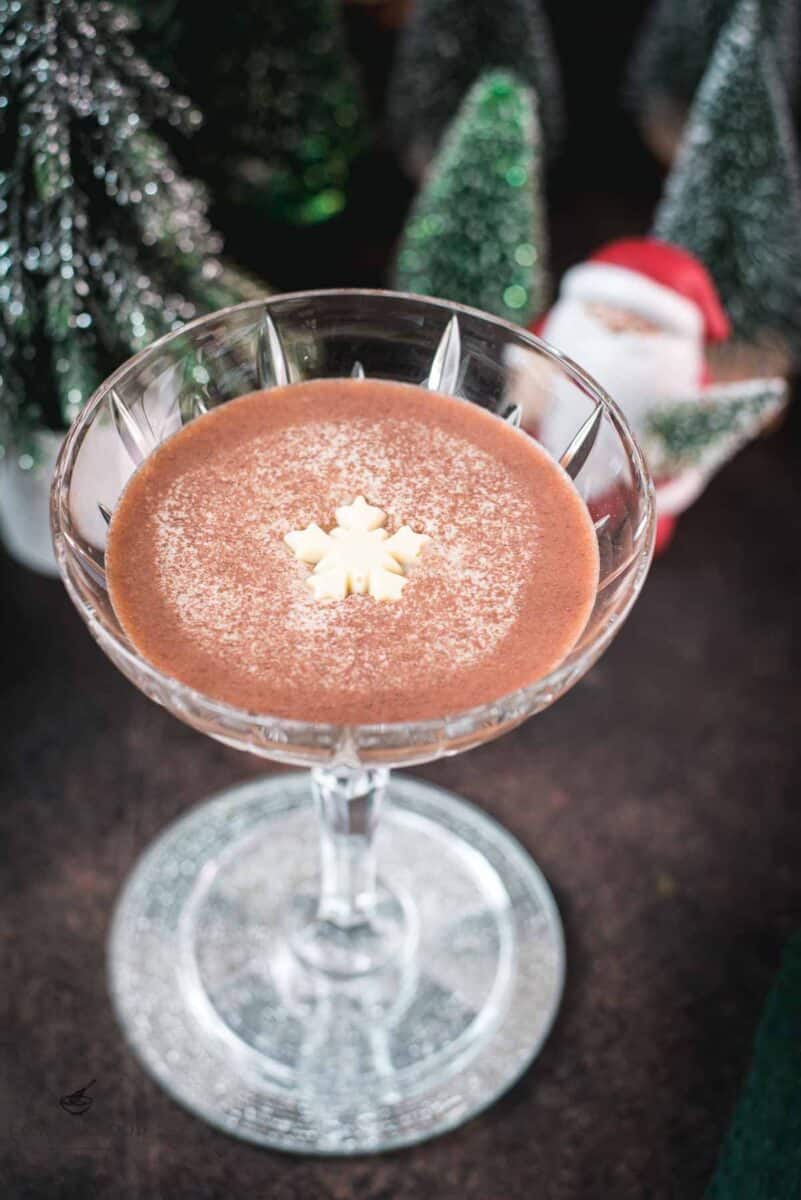 Festive chocolate martini with a white chocolate snowflake garnish, placed on mirrored coaster next to a Santa clause figurine.