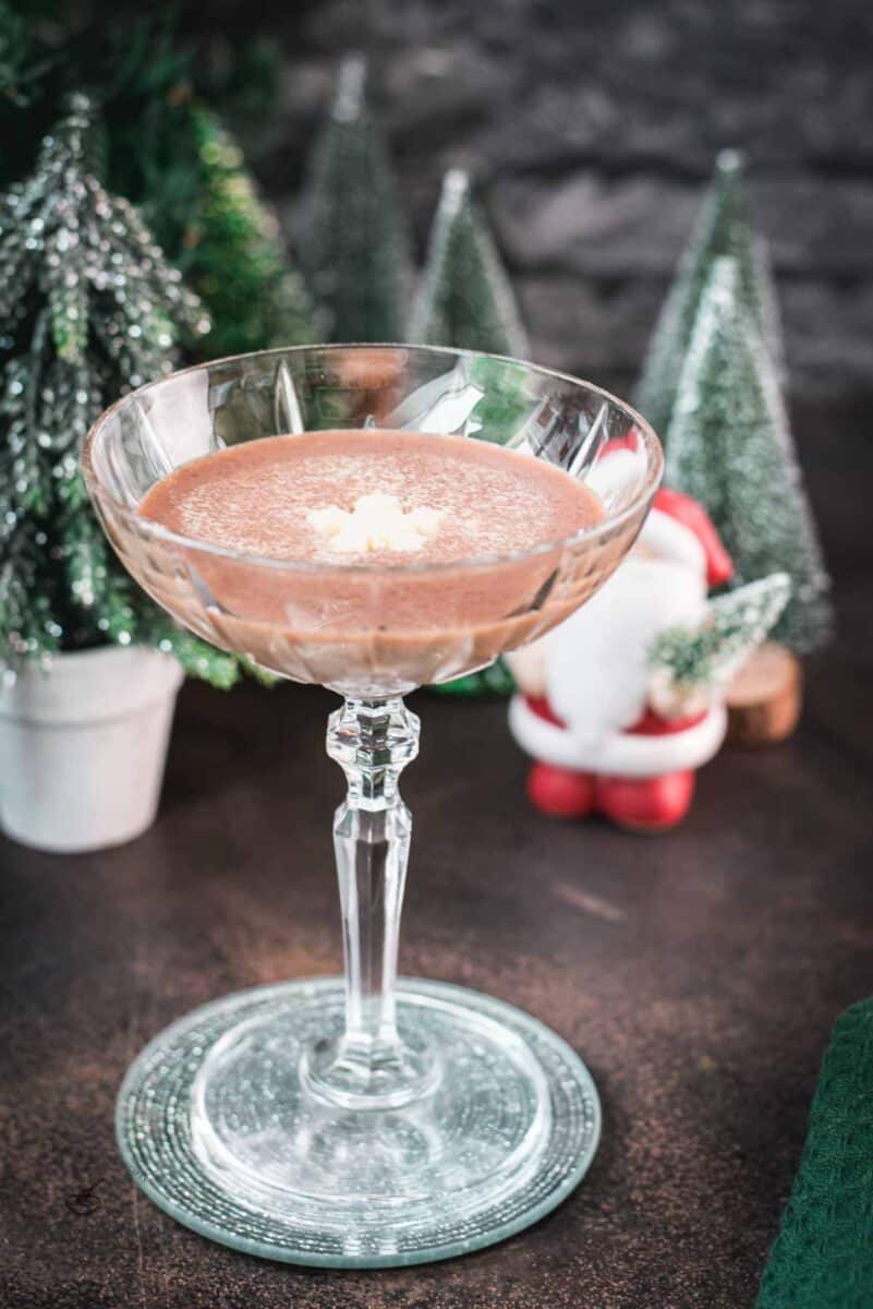 Festive chocolate martini with a white chocolate snowflake garnish, placed on mirrored coaster next to a Santa clause figurine.