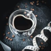Heart-shaped coffee mug filled with black coffee, next to measuring tape on dark ground.