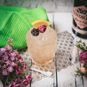 Fancy bramble cocktail in gorgeous glass, placed next to flowers and a white spoon. Garnished with a blackberry, raspberry, and a lemon wedge.