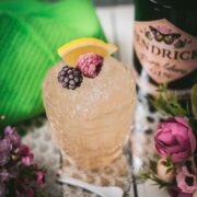 Fancy bramble cocktail in gorgeous glass, placed next to flowers and a white spoon. Garnished with a blackberry, raspberry, and a lemon wedge.