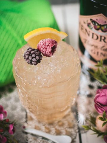 Fancy bramble cocktail in gorgeous glass, placed next to flowers and a white spoon. Garnished with a blackberry, raspberry, and a lemon wedge.