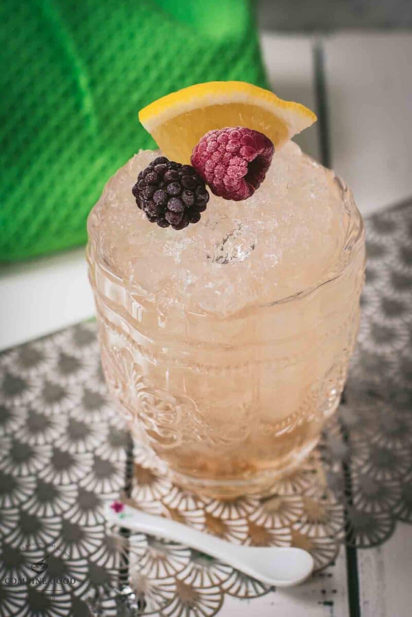 Fancy bramble cocktail in gorgeous glass, placed next to flowers and a white spoon. Garnished with a blackberry, raspberry, and a lemon wedge.