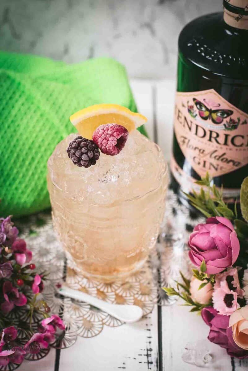 Fancy bramble cocktail in gorgeous glass, placed next to flowers and a white spoon. Garnished with a blackberry, raspberry, and a lemon wedge.