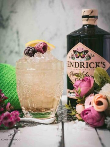 Fancy bramble cocktail in gorgeous glass, placed next to flowers and a white spoon. Garnished with a blackberry, raspberry, and a lemon wedge.