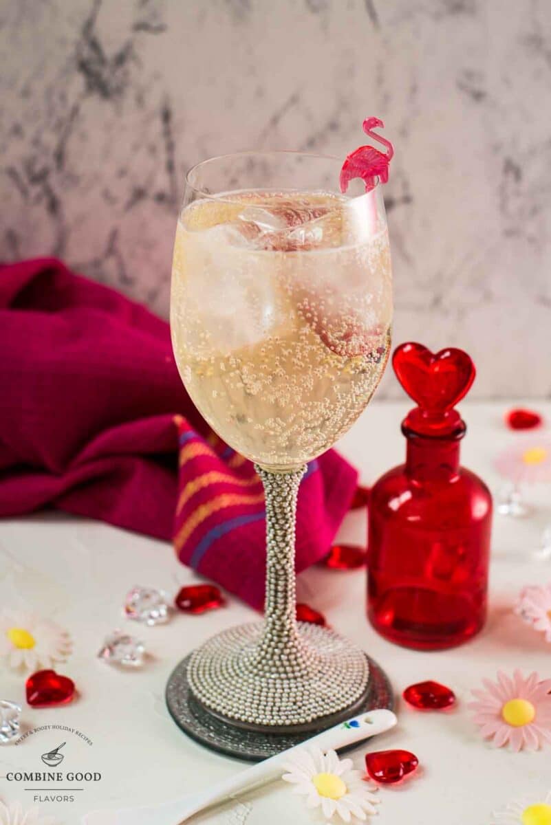 Valentines gin cocktail, garnished with a gorgeous flamingo cocktail pick, served in wine glass. Placed on a mirrored coaster next to a heart shaped flask.