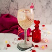 Valentines gin cocktail, garnished with a gorgeous flamingo cocktail pick, served in wine glass. Placed on a mirrored coaster next to a heart shaped flask.