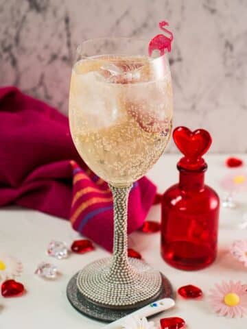 Valentines gin cocktail, garnished with a gorgeous flamingo cocktail pick, served in wine glass. Placed on a mirrored coaster next to a heart shaped flask.