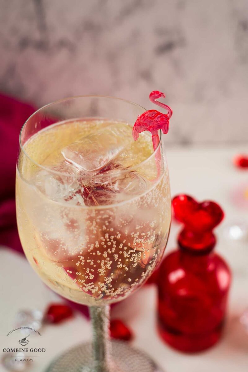 Valentines gin cocktail, garnished with a gorgeous flamingo cocktail pick, served in wine glass. Placed on a mirrored coaster next to a heart shaped flask.