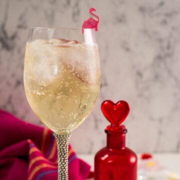 Valentines gin cocktail, garnished with a gorgeous flamingo cocktail pick, served in wine glass. Placed on a mirrored coaster next to a heart shaped flask.