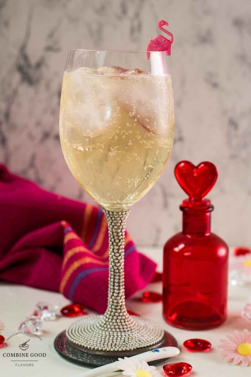 Valentines gin cocktail, garnished with a gorgeous flamingo cocktail pick, served in wine glass. Placed on a mirrored coaster next to a heart shaped flask.