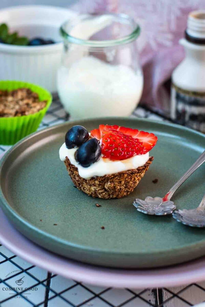 Cute little granola cup, filled with yogurt and topped with fresh strawberries, blue grapes, and drizzled with maple syrup. Placed on dark green plate.