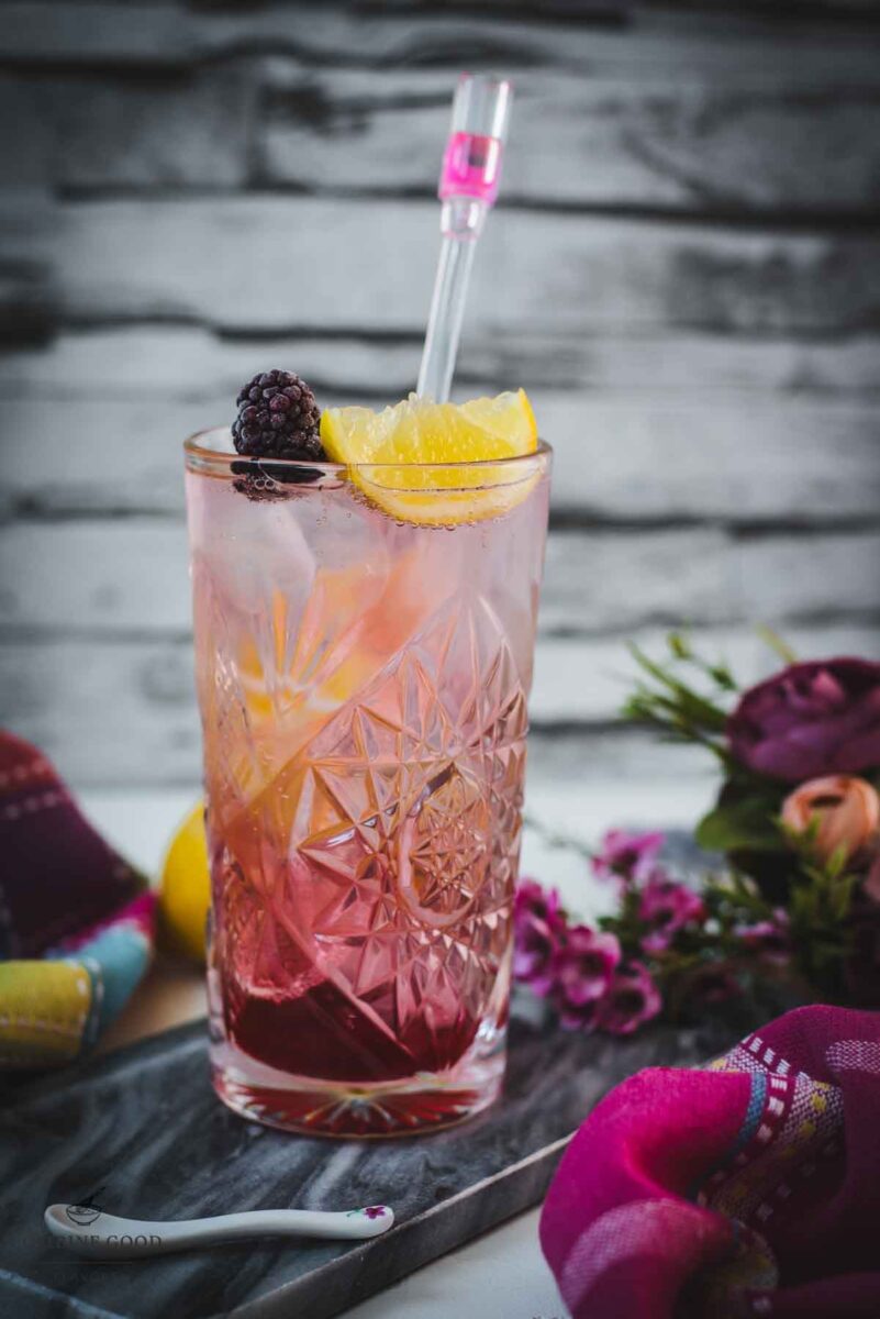 Refreshing blackberry raspberry gin fizz, garnished with a lemon wedge and a blackberry, placed on marbled plate.