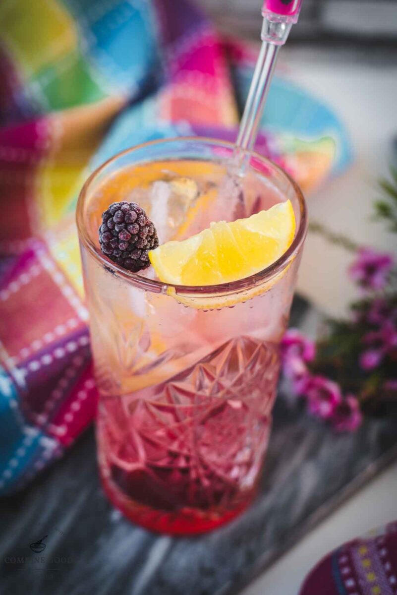 Refreshing blackberry raspberry gin fizz, garnished with a lemon wedge and a blackberry, placed on marbled plate.