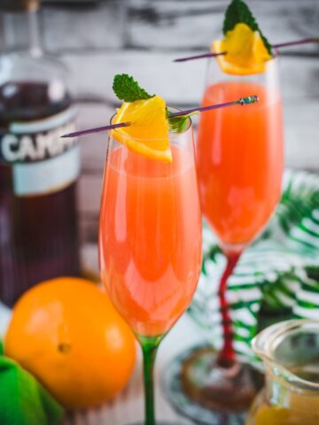 Two champagne glasses filled with vibrant Campari Mimosa. Garnished with an Orange slice and mint leaf on a cocktail skewer.
