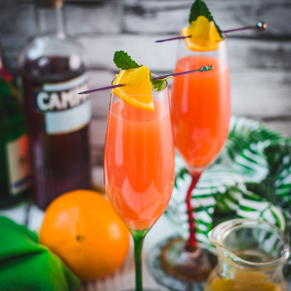 Two champagne glasses filled with vibrant Campari Mimosa. Garnished with an Orange slice and mint leaf on a cocktail skewer.
