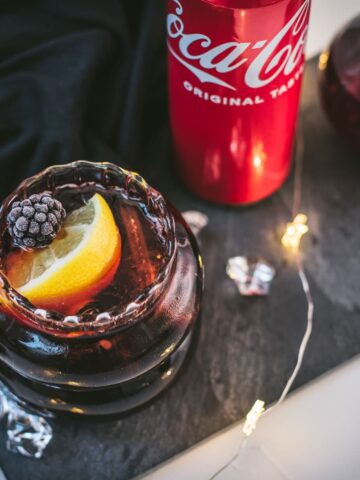 Cute glass filled with a delicious chambord coke cocktail, placed on black slate plate and garnished with a lemon wedge and one frozen blackberry.