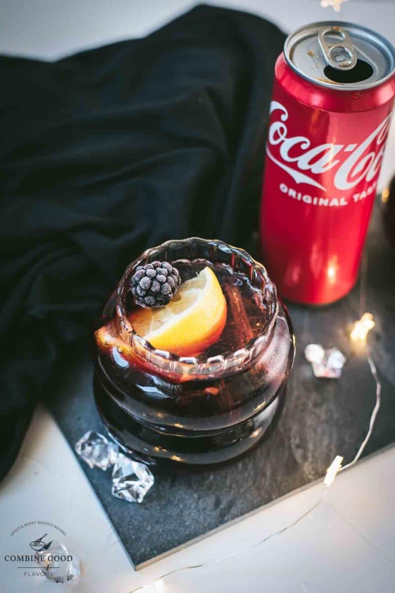 Cute glass filled with a delicious chambord coke cocktail, placed on black slate plate and garnished with a lemon wedge and one frozen blackberry.