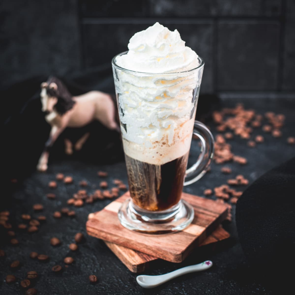Beautiful tall glass filled with strong coffee and topped with whipped cream. Placed on brown coasters and pretty horse in the background. The Einspänner, a Viennese classic!