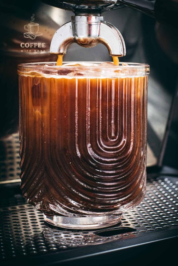 Brewing process of iced Americano into a iced and cold water filled serving glass, using an espresso machine.