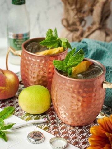 Delicious peach Moscow mule, served in copper mugs, garnished with peach slices and mint.