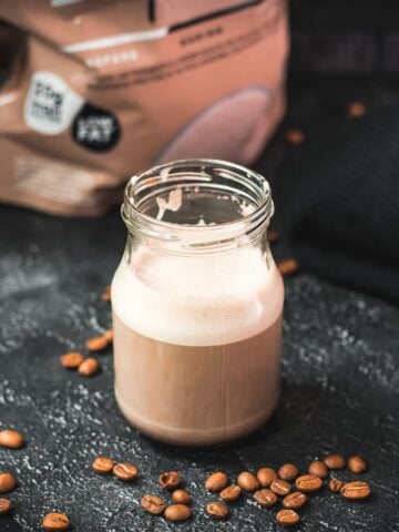 Overhead view of hot protein coffee in a glass yogurt jar on a dark surface, surrounded by coffee beans and a visible whey protein pouch in the background.