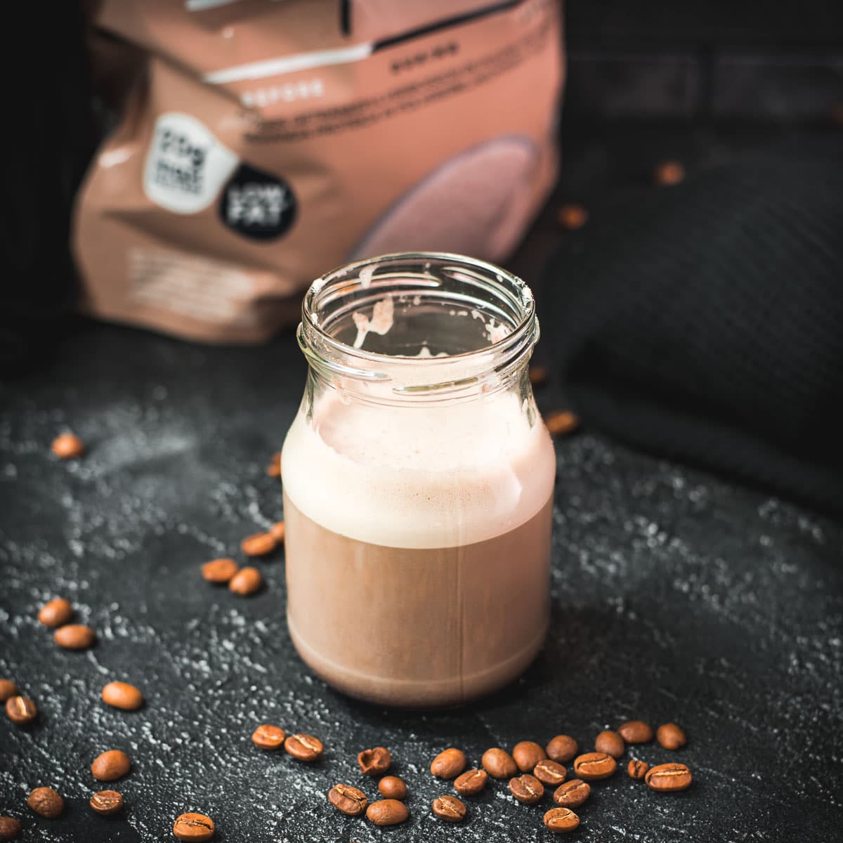 Overhead view of hot protein coffee in a glass yogurt jar on a dark surface, surrounded by coffee beans and a visible whey protein pouch in the background.