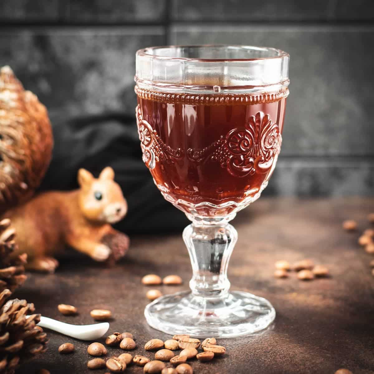 Gorgeous glass filled with maple syrup coffee, placed on dark brown ground, next to squirrel figure.