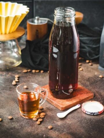 Amazing coffee syrup, in small glass next to a preserving bottle filled with coffee syrup, placed on brown ground next to coffee beans.